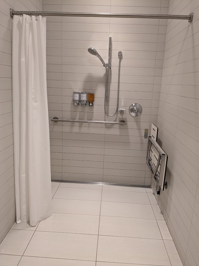 A shower area, with curtain on the left, bench on the right, and shower head and shampoo on the back wall.